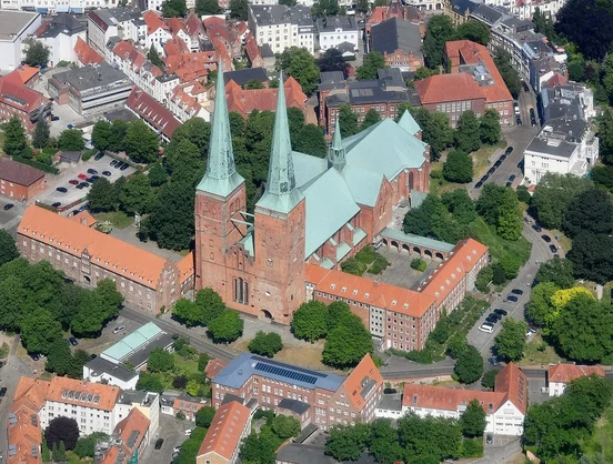 The image shows an aerial view of a historic building, most likely a cathedral or church, characterized by its tall spires and distinct architectural features. Surrounding it are other buildings, trees, and streets, suggesting it is located in a developed area with urban elements. The layout indicates a mixture of green spaces and residential or commercial structures. The red roofs are a notable architectural detail in the area.

Image Credits: Wikimedia / Carsten Steger / CC BY-SA 4.0