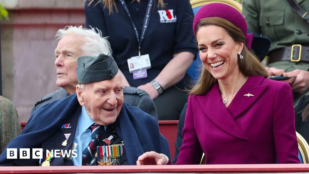A veteran smiles while wearing military uniform and badges while Catherine, Princess of Wales  laughs sitting next to him wearing a maroon suit and hat, while they watch the procession arrive at Buckingham Palace on Monday.