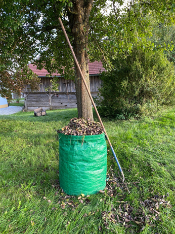 ein grüner großer Sack gefüllt bis oben hin mit Blättern, an dem ein Rechen lehnt 