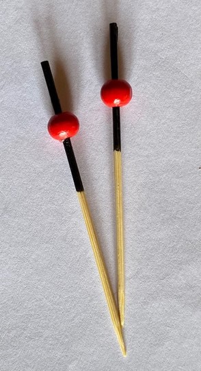 zwei kleine Spießchen liegen nebeneinander. nDie obere Hälfte ist schwarz und in der Mitte vom Schwarz sitzt eine leuchtend rote kleine Kugel 