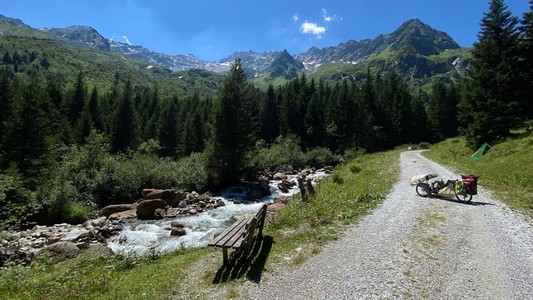 auf einem geschotterten Weg an einem Bach mit Aussichtsbank steht ein Liegedreirad nim Hintergrund hohe Berge