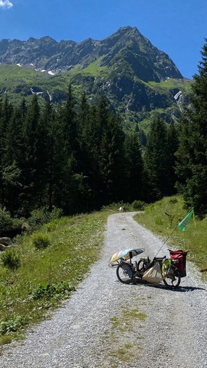 ein Liegedreirad steht auf einem geschotterten Weg in der Sonne in die hohen Berge hinein 