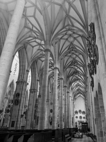 ein schwarweiß Foto eines der Seitenschiffe vom Ulmer Münster noberhalb von vielen runden Säulen und einer zweiten Reihe eckiger Säulen ein Muster von helldunkel Verbindungen im Deckengewölbe