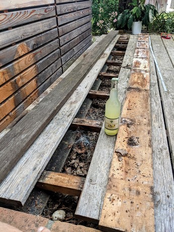 eine Holzterrasse mit Hochbeet daneben. Eine Lücke und zwei Bretter liegen links und rechts daneben. Das alte morsche Brett, und ein noch zu langes Ersatzbrett. Eine Flasche Zitronenlimonade steht auch da.