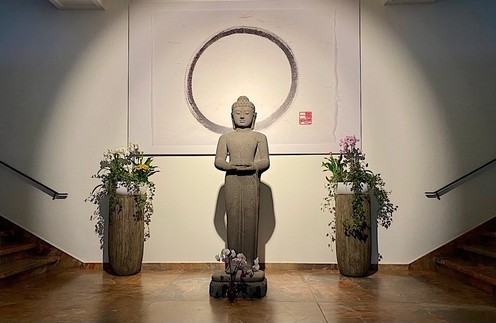 eine graue stehende Buddhastatue mit dekorativen Pflanzen steht vor einer Kalligrafie eines fast ganz geschlossenen Kreises und ist beleuchtet. Links und rechts sind Treppen erkennbar 