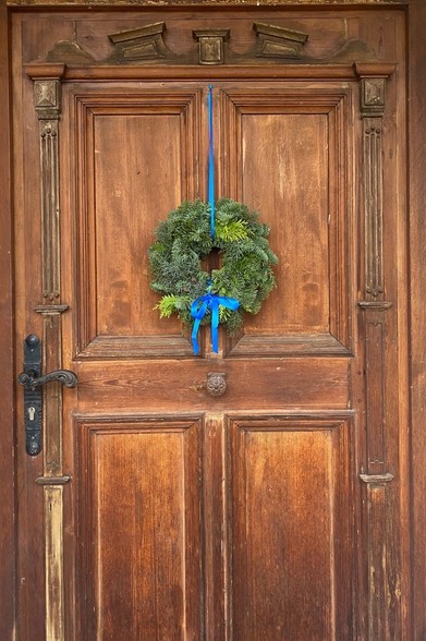 an einer hölzernen Haustür mit Kassetten hängt ein grüner Kranz an einem blauen Band mit blauer Schleife 