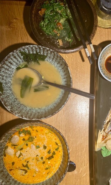 drei Schalen auf einem Tisch von oben fotografiert mit Seegrassalat, Misosuppe und Kokossuppe mit Koriander und ein Teil einer langen Sushiplatte auf der hellgrüner Wasabi erkennbar ist