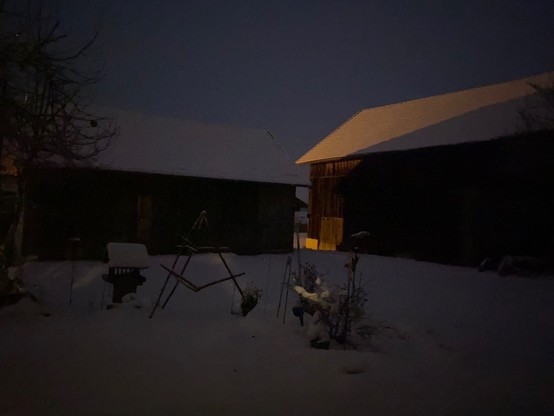 der Innenhof eines ehemaligen Bauernhofs ist schneeweiß. Eine kleine und eine große Scheune stehen im rechten Winkel. Das Dach der großen Scheune ist teilweise orange beleuchtet. 