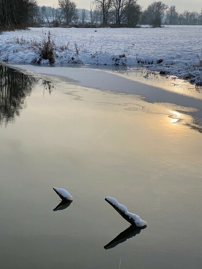 eine verschneite Landschaft mit einem spiegelnden kleinen Fluss in dem eine fahle Sonne zu erahnen ist und zwei schiefe verschneite Holzpflöcke ragen aus dem Wasser und werfen Schatten 