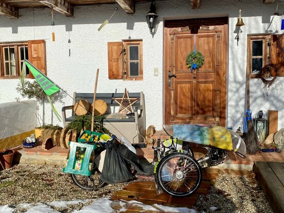 vor der Haustür eines alten Bauernhauses steht ein mit 2 Getränkekisten beladenes Liegedreirad mit Wind- und Regenschutz und Fahne hinten 