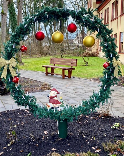ein großer Kreis geschmückt mit Fichtenzweigen und roten und golden Weihnachtskugeln und dahinter ein Weg mit Holzbank und grünem Rasen 