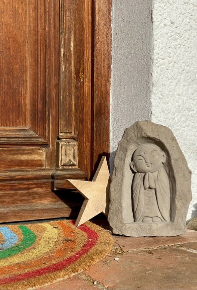 vor einer hölzernen Haustür steht vor einer weißen Hauswand ein kleiner grauer „Kindmönch“ mit gefalteten Händen in einer angedeuteten Nische. Dahinter lugt ein Holzstern hervor. 