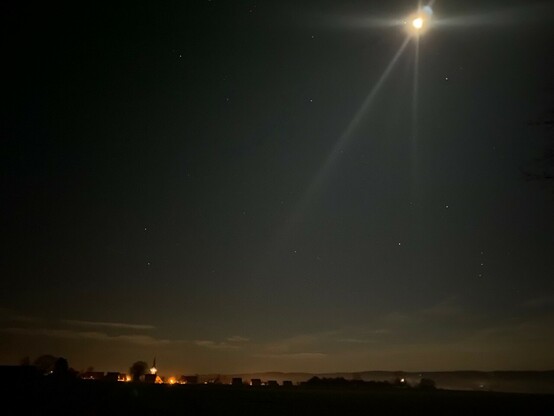 der Mond an einem fahlen Himmel mit ein paar Sternen und am Horizont ein paar wenige orange Lichter eines Dorfes