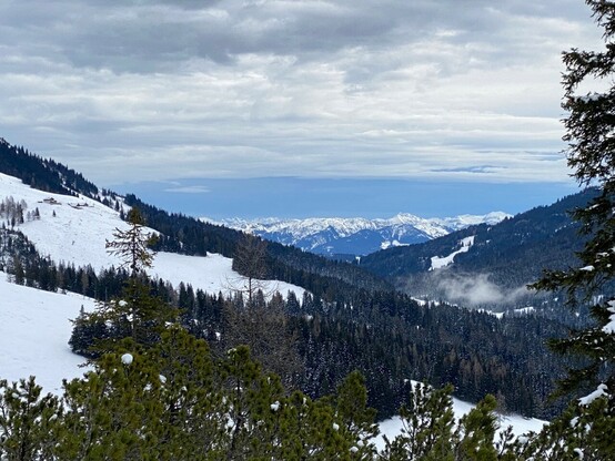 Ausblick ins Tal und ein entfernt liegende verschneite Bergendunkle Wolken, helle Wolken und keine Wolken am Himmel 