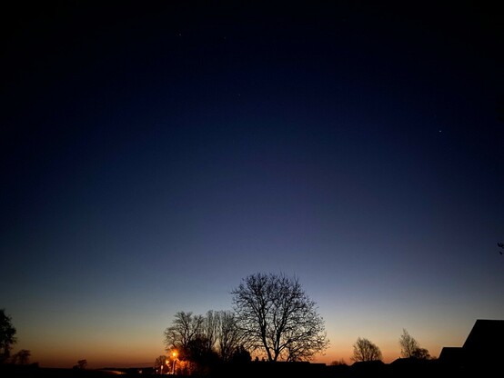 Dämmerung mit erstem Orange am Horizont und noch erkennbar Sternen am dunklen Himmel und einer Straßenlaterne 