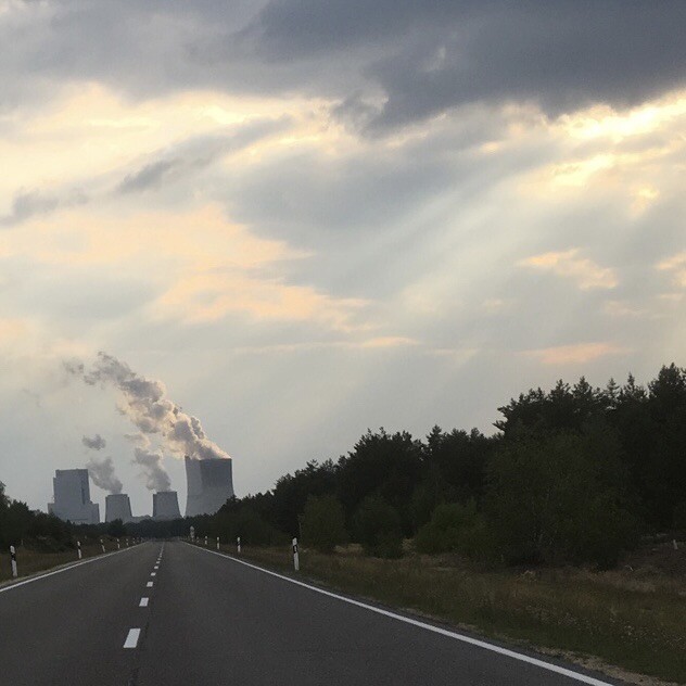 Kraftwerk am Ende einer langen geraden Straße. Die Kühlturmwolken wehen leicht nach links. Von rechts oben dringen Sonnenstrahlen durch echte Wolken.