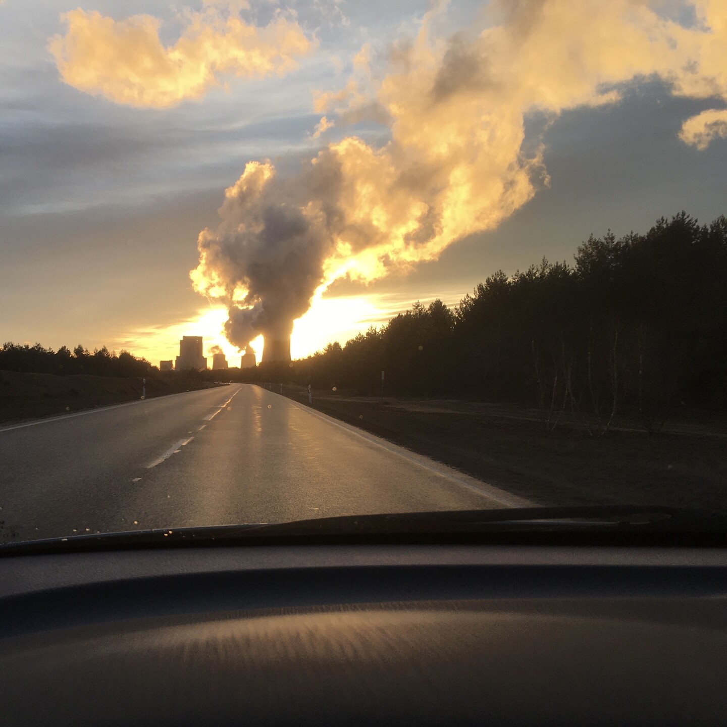 Kraftwerk am Ende einer langen geraden Straße. Diese ist feucht, das Kraftwerk spiegelt sich darauf. Die Sonne steht direkt hinter den dicken Kühlturmwolken.