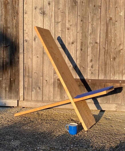 ein einfacher Steckstuhl mit einem blauen Sitzkissen und einer blauen Tasse stehen vor einem hölzernen Scheunentor in der Sonne