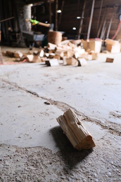 auf einem Betonboden liegt ein einzelner Holzscheit. Im Hintergrund sind unscharf ein Hackstock mit Spaltaxt und rundherum gespaltenem Holz zu sehen 