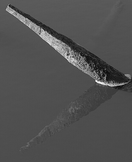 Schwarzweiß nnein spitzer Pfahl ragt mit weniger als 45 Grad aus dem glatten Wasser und spiegelt sich
