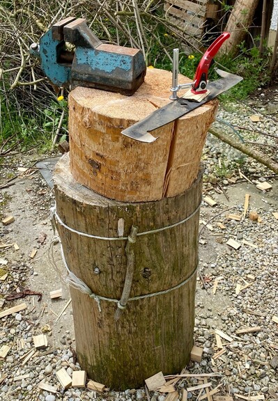 auf einem Hackstock steht ein weiterer Baumanschnitt mit ähnlichem Durchmesser, auf dem ein Schraubstock befestigt ist.nEine Klemme steckt in einem Loch und hält das Rasenmähermesser flach auf dem Baumstück fest