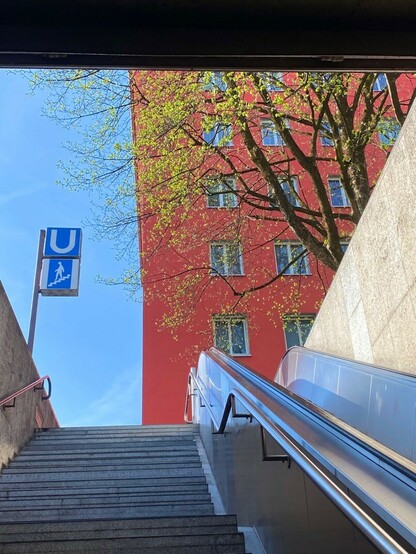 U-Bahnausgang mit Treppe hoch und blauem Himmel und einem roten Haus und kleinen grünen Blättern an einem Baum 