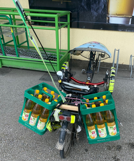 ein Liegedreirad steht vor einem Radlständer und es hängen links und rechts je eine volle 12er Getränkekiste am Gepäckträger 