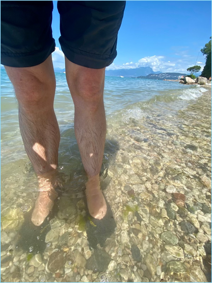 Füße mit Barfußschuhen stehen im Wasser und um Hintergrund sind Berge zu erkennen 