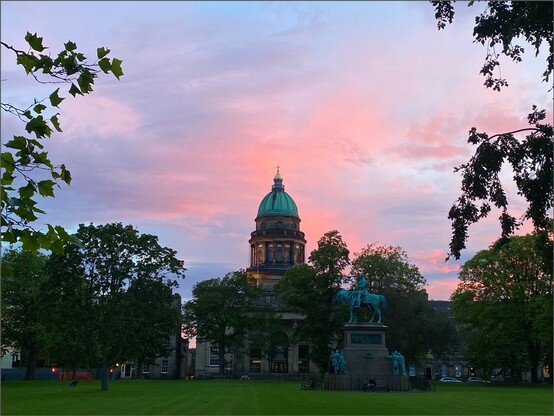 nach Sonnenuntergang ein paar rosa Wolken über einem Gebäude mit halbkugelförmigen kupfergrünem Dach. Auf dem Rasen davor steht eine kupfergrüne Reiterstatue