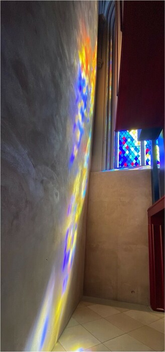 in einer Kirche scheint die Sonne durch ein farbiges Fenster und erzeugt ein buntes Farbmuster auf einer beigen Wand. Rechts davon Teile der rötlichbraunen Orgel 