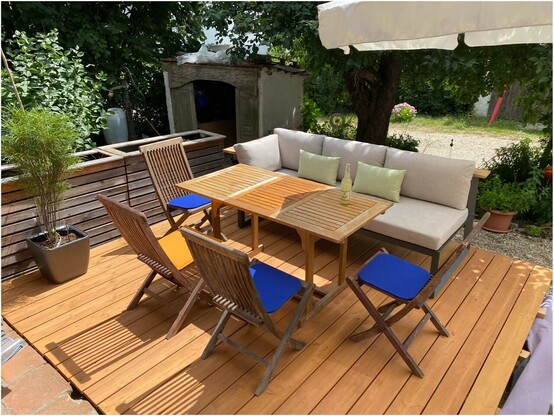 auf einer Holzterrasse stehen 4 Holzstühle mit 3 blauen und einem orangen Sitzkissen an einem Holztisch. An einer Längsseite steht eine Couch mit beigen Sitzkissen