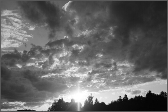 Schwarzweiß Foto in Richtung der am Horizont verschwindenden Sonne mit Wolken. Die Sonne strahlt richtig unter den Wolken durch 