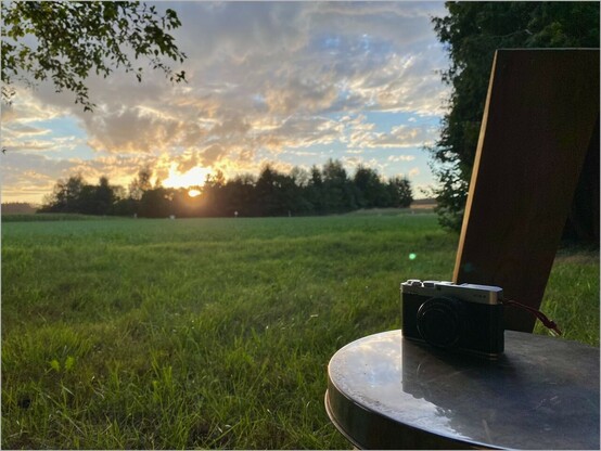 Blick auf farbige Wolken eines Sonnenuntergangs unter einem Baum heraus. Im Vordergrund steht eine Kamera auf einem Edelstahl Tablett. Die Lehne von einem Steckstuhl am rechten Rand