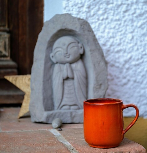 ein leuchtend oranger Kaffeebecher steht vorm Haus und ein kleiner Jizu Buddha steht dahinter. Ein kleiner Holzstern ist teilweise zu erkennen