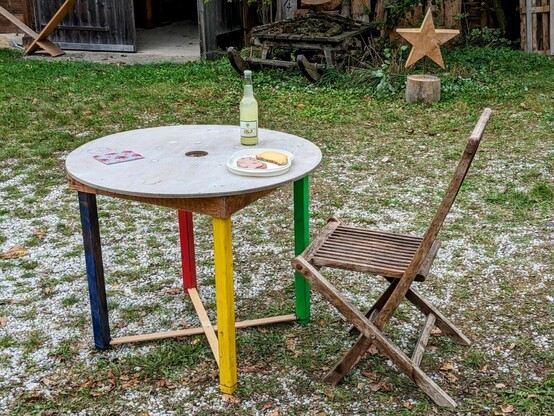 ein runder Tisch mit bunten Beinen und ein hölzerner Klappstuhl stehen auf grasdurchwachsenem Kies im Innenhof. Eine Limoflasche und 2 Brote auf dem Tisch