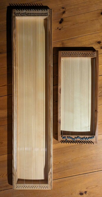 zwei Monochorde liegen auf dem Holzboden. Das längere ist doppelt so lang, aber genauso breit und hat auch gleich viele Saiten
