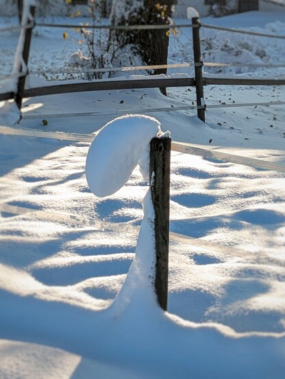 Die Schneehaube auf einem Zaunpfahl ist vom Wind nach links gekippt und der Halt sieht sehr fragil aus.
