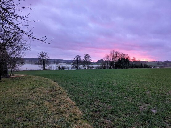 rechts am wolkigen Himmel ein paar rosa Flecken von einem Sonnenuntergang.nIm Vordergrund Wiese und grünes Feld. Dahinter eine große Wasserfläche (die da normalerweise nicht ist).