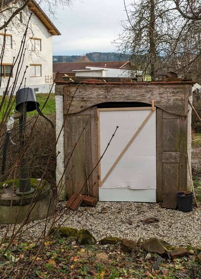 ein kleines weißes Hüttchen mit unvollständiger Holzfront hat eine unvollständige weiße Tür bekommen.nKies vor dem Hüttchen.