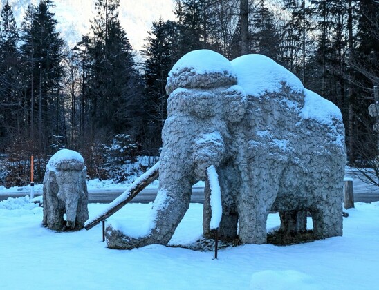 zwei aus Stein gehauene Mammuts (Baby und Erwachsenes) stehen im Schnee. Dahinter lockere Nadelbäume