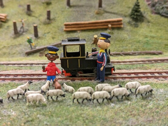 Jim Knopf und Lokomotivführer aus der Augsburger Puppenkiste stehen mit ihrer Lokomotive bei Schafen auf einer Wiese
