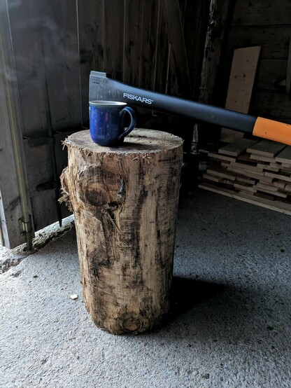 eine blaue Tasse steht auf einem Baumanschnitt ohne Rinde, und eine Axt mit schwarzorangem Griff steckt im Holz