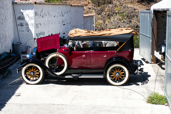 Ein schöner alter weinrot lackierter Buick mit Faltdach, Weißwangenreifen und Holzspeichen steht mit hochgeklappter Motorhaube vor einer Garage. 