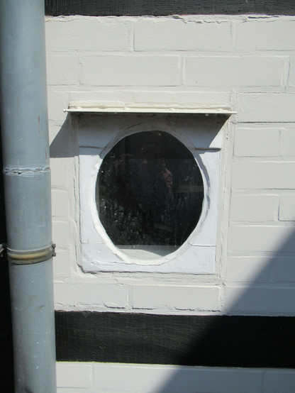 Ein ovales, kleines Fenster in einem Fachwerkhaus mit schwarz gestrichenen Balken und weiß getünchtem Klinker. Links ein Fallrohr.