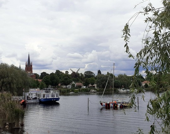 Bild von der Havel in Werder mit Booten auf dem Wasser, viele Pflanzen, einer Windmühle und einer Kirche. Der Himmel ist voller Wolken.