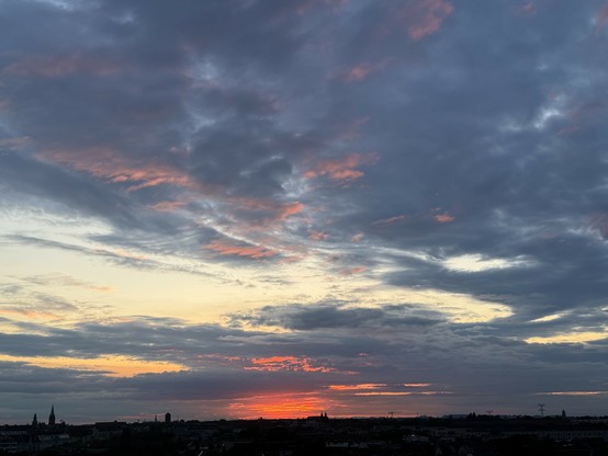 Sunset with a mostly cloudy sky, displaying various shades of orange, pink, and gray. The city skyline is visible in the lower part of the image.
