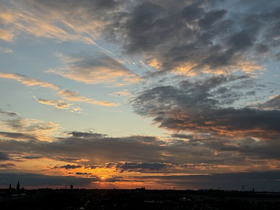 A panoramic view of a sunset with a city skyline in the foreground and dramatic clouds illuminated by the setting sun.