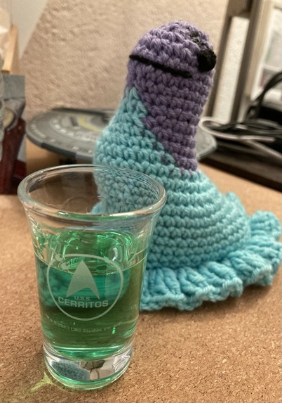 A Cerritos shot glass with green woodruff liqueur, in the background a crocheted Murf and USS Cerritos model.