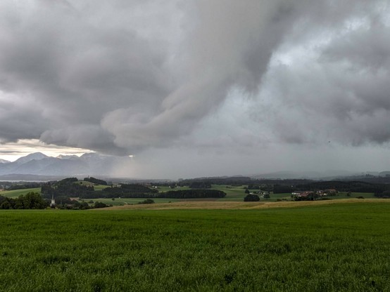 dunkle Wolken am Himmel, von rechts schieben sich Wolken rein, die bis zum Boden reichen und schon deutlich mehr Berge sind hinter den Wolken verschwunden ngrüne Wiese im Vordergrund und Chiemgauer Berge im Hintergrund