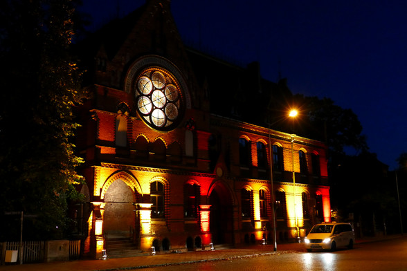 Ein Gebäude im Stil des Historismus wird mit rot und gelb strahlenden Scheinwerfern steil von unten angestrahlt. Die Fensterhöhlungen bleiben dabei im Schatten.